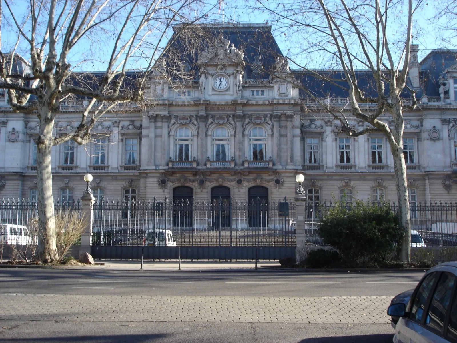 Façade de la préfecture du Rhône à Lyon, avec son horloge et ses grilles, derrière des platanes